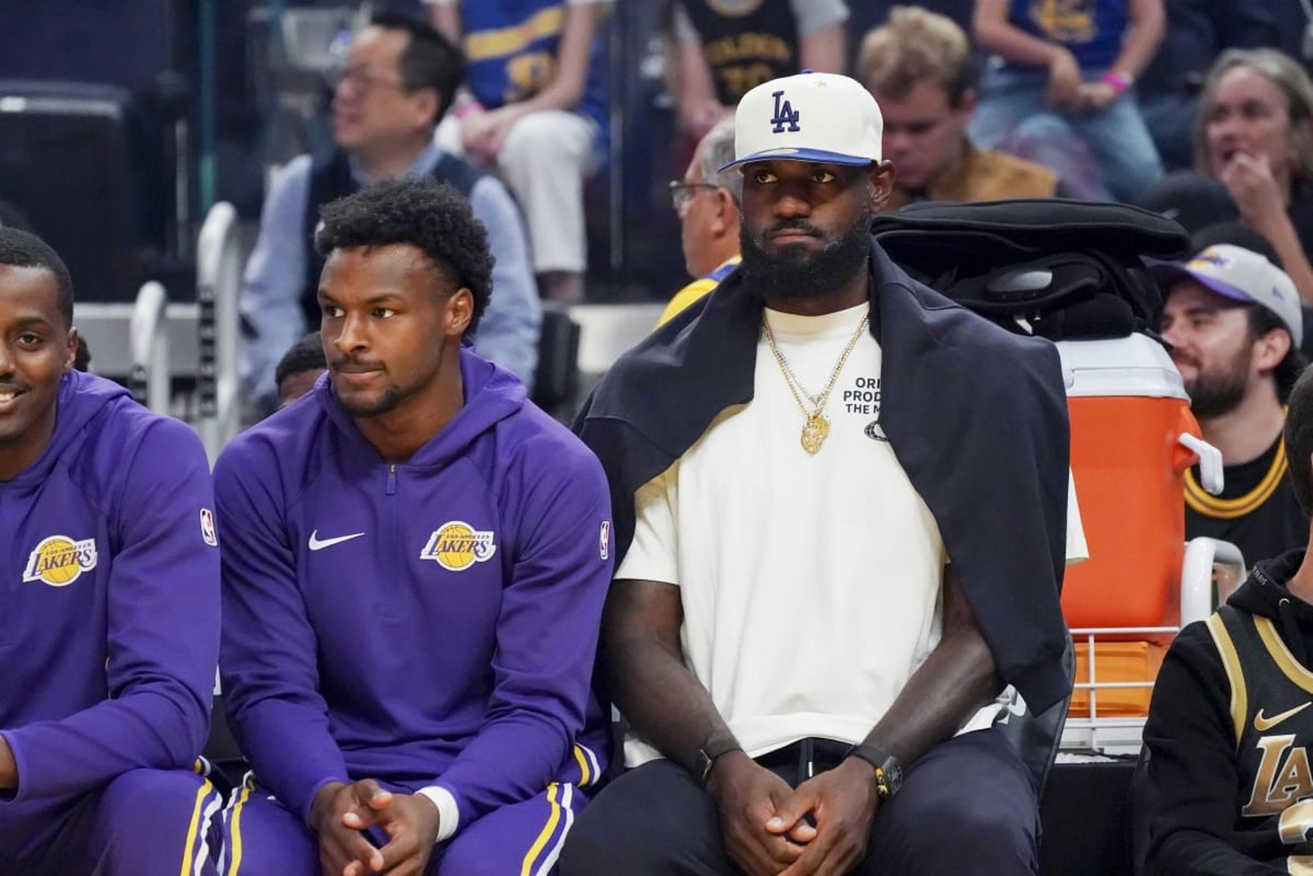 Oct 5, 2025; San Francisco, California, USA; Los Angeles Lakers guard Bronny James (9) and forward LeBron James (23), wearing a Los Angeles Dodgers cap, watch the action against the Golden State Warriors in the first quarter at Chase Center. Mandatory Credit: David Gonzales-Imagn Images